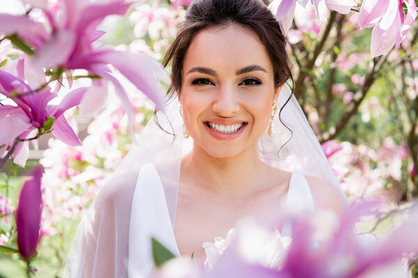 Счастливая невеста в вуали улыбается в камеру возле цветущей магнолии 