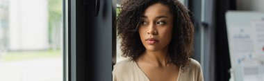 portrait of thoughtful african american woman standing near window in modern office, banner