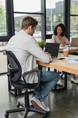 interracial colleagues sitting at desks and working with laptops in modern open space