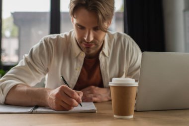 focused young man sitting at desk near paper cup and laptop and writing with pen in notebook in modern office