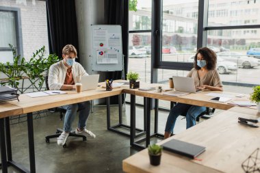 young interracial colleagues in medical masks working with laptops in modern office
