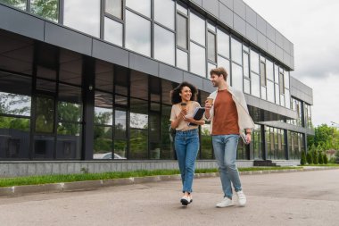 smiling young interracial couple with paper coffee cups and cellphones in hands walking on street near modern building