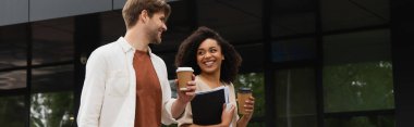 smiling young interracial couple with paper cups looking at each other and walking near building, banner