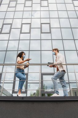 low angle view of young interracial couple in medical masks and casual clothes talking near glass building on street