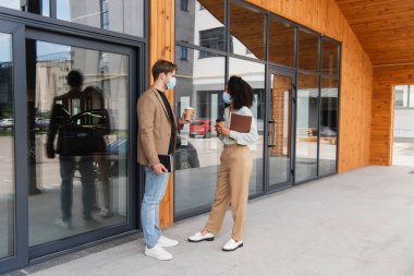 young interracial colleagues in medical masks and business casual clothes talking with paper cups in hands near building