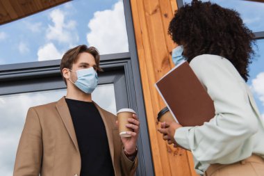 low angle view of young interracial colleagues in medical masks talking with paper cups in hands near building