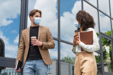 young interracial colleagues in medical masks walking with coffee paper cups and documents near modern glass building