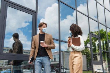 young interracial colleagues in medical masks walking with coffee paper cups and documents near modern building