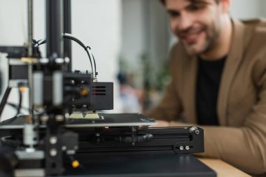 3D printer producing plastic model near blurred young smiling man in modern office