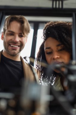 cheerful young interracial colleagues looking at blurred 3D printer in modern office