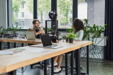 smiling interracial colleagues sitting with cellphones in hands near 3D printer and laptops in modern office