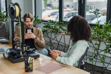 young interracial colleagues sitting with cellphones in hands near 3D printer in modern office