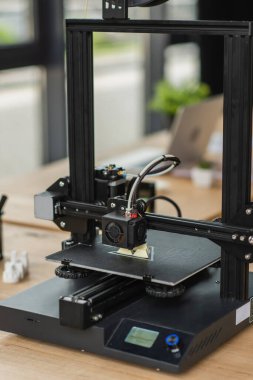 3D printer creating plastic model near blurred figures and laptop in modern office