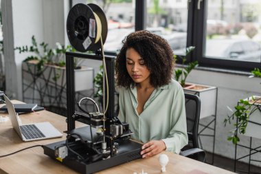 serious young designer working with 3D printer near laptop and produced plastic models on table in modern office