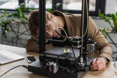 concentrated young designer looking at 3D printer producing plastic figure near laptop and cellphone in modern office