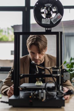 focused young designer looking at 3D printer producing model in modern office
