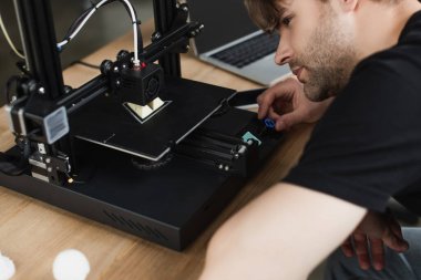 concentrated young designer looking at 3D printer producing plastic figure in modern office