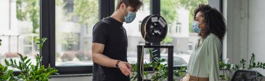 interracial colleagues in medical masks standing near 3D printer in modern office, banner