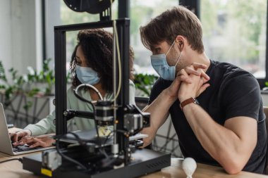 interracial colleagues in medical masks working with 3D printer and laptop in modern office