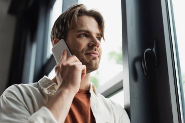 positive young man in beige shirt talking on cellphone near window in modern office