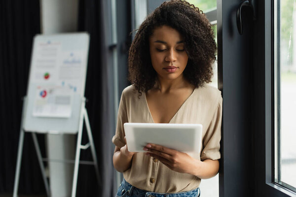 african american woman in casual clothes standing near window and typing on digital tablet in modern office