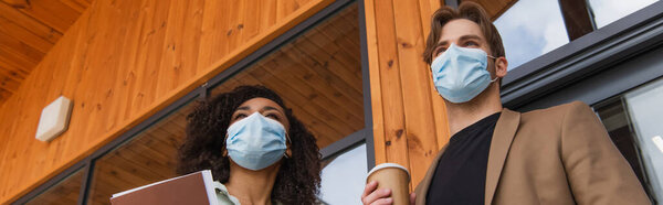 низкий угол обзора молодых межрасовых коллег в медицинских масках, стоящих на улице возле здания, баннер