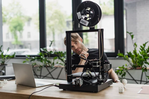 young designer looking at 3D printer on table with laptop in office