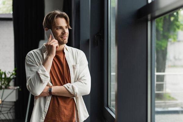 positive young man in beige shirt talking on cellphone near window in modern office