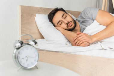 Genç adam komodinin üzerinde bulanık çalar saatin yanında uyuyor. 