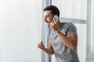 Side view of excited man showing yes gesture while talking on smartphone near window  clipart