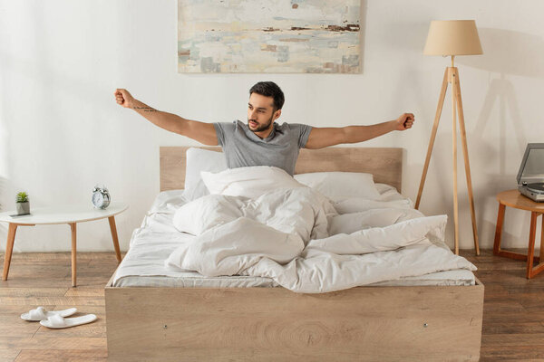 Bearded man stretching while sitting on bed in morning 