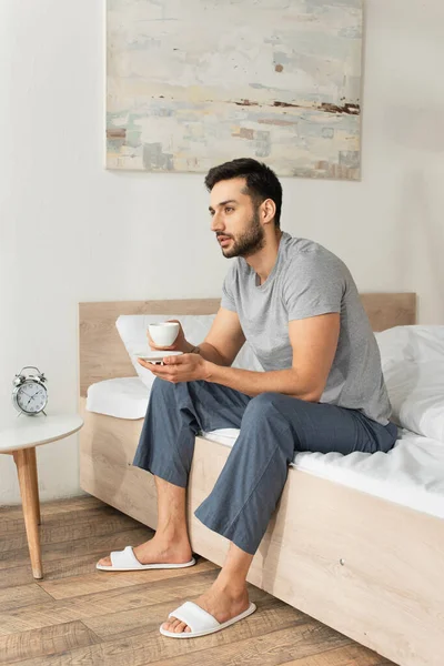 Young man in pajama and slippers holding coffee cup on bed