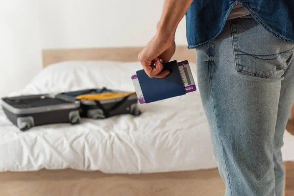 ベッドの上でぼやけたスーツケースの近くにパスポートと航空券を保持している男の作物ビュー — ストック写真