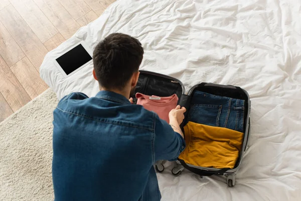 ベッドの上でデジタルタブレットの近くのスーツケースに服を着る男のオーバーヘッドビュー — ストック写真