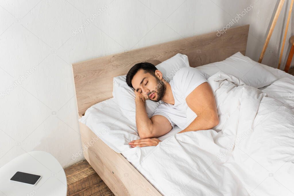 Bearded man sleeping on bed near smartphone with blank screen on bedside table