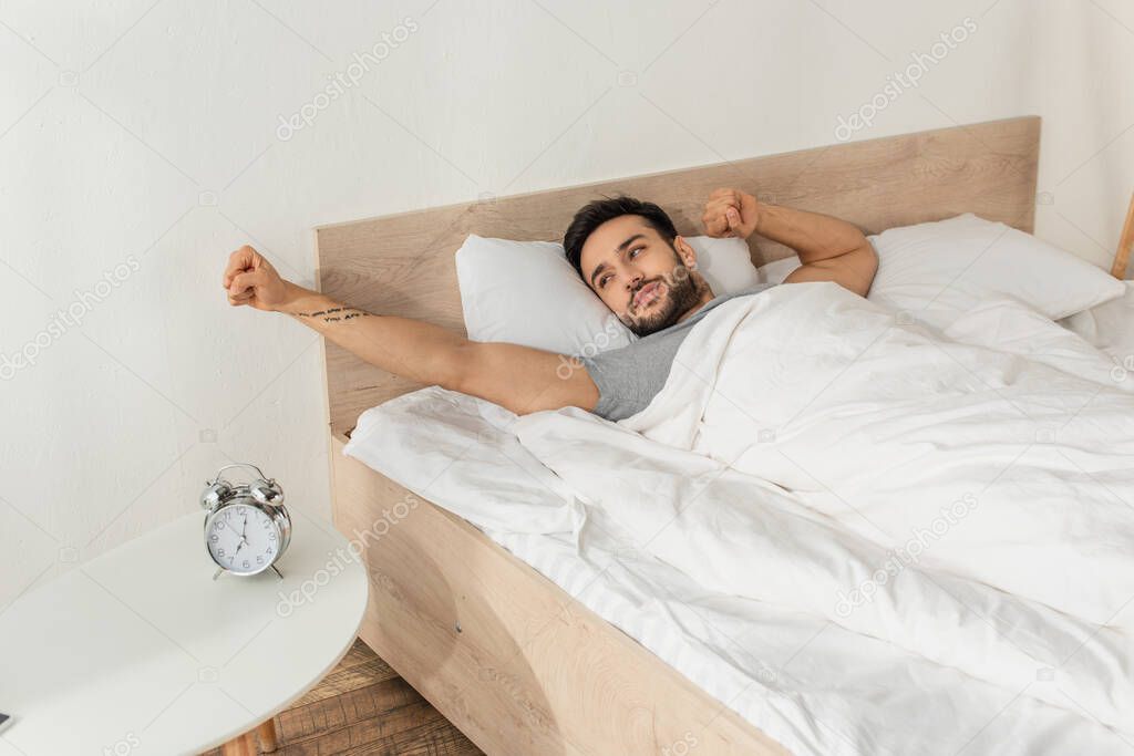 Young man stretching and looking at alarm clock near bed 