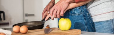 Cropped view of young coupe cooking pancake near eggs and apple, banner  clipart