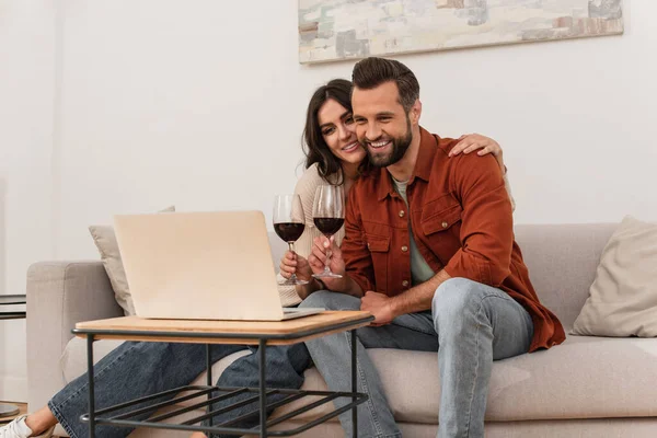 Mulher Sorridente Abraçando Namorado Com Copo Vinho Perto Laptop Casa — Fotografia de Stock