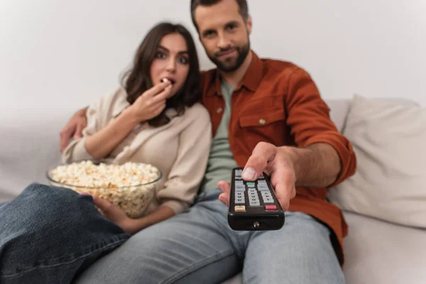 Remote controller in hand of man near girlfriend with popcorn on couch