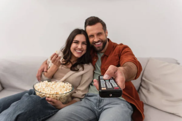 Remote controller in hand of blurred man near cheerful girlfriend with popcorn