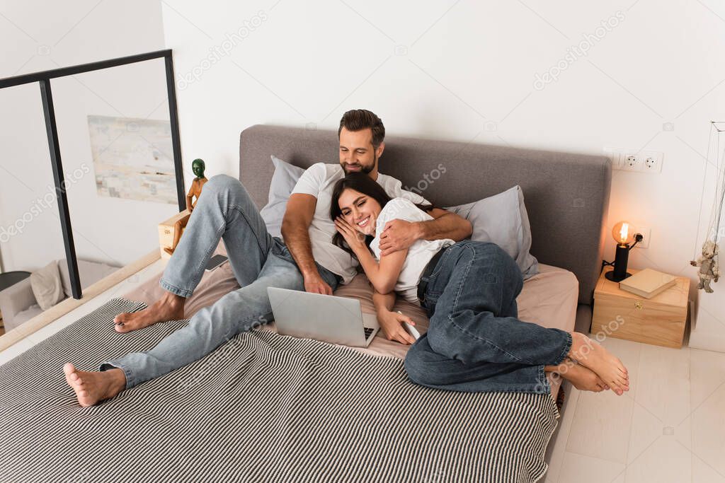 Man hugging girlfriend wit smartphone near laptop on bed 