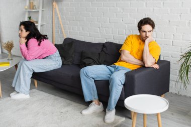 disappointed young man with hand near face sitting on couch near girlfriend in living room clipart