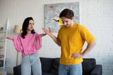 angry young woman standing with hands in air near boyfriend with refuse gesture in living room clipart