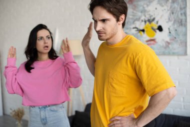 angry young woman shouting with hands in air near boyfriend with refuse gesture in living room clipart