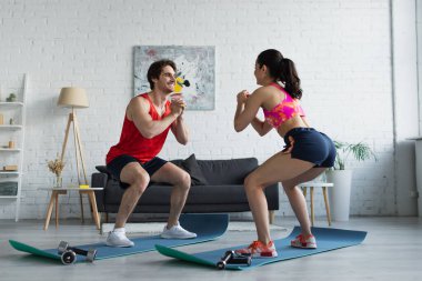 Spor kıyafetleri içinde gülümseyen genç bir çift evdeki spor minderlerine çömeliyor.