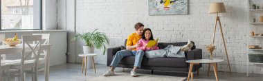 smiling young man hugging girlfriend lying on couch with book in hands in living room, banner clipart