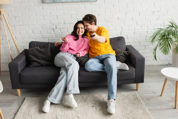 Smiling Young Man Pointing Remote Controller Camera Hugging Girlfriend Home — Stock Photo, Image