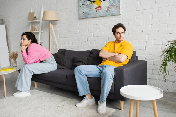 disappointed young man with crossed arms sitting on couch near girlfriend with hands near face in living room