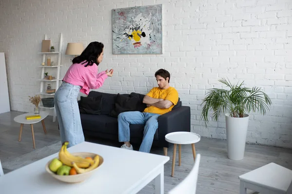 Vred Ung Kvinde Gesturing Med Hænderne Nær Kæreste Sidder Sofaen - Stock-foto