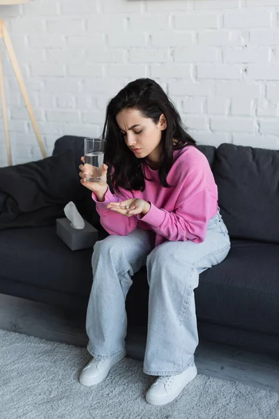 Mujer Joven Enferma Sentada Sofá Con Vaso Agua Mirando Píldoras —  Fotos de Stock